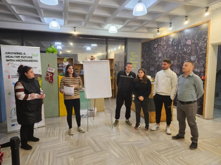 A woman holding a sheet of paper speaks to a small group standing beside a flipchart during the microgreens training course in Athens