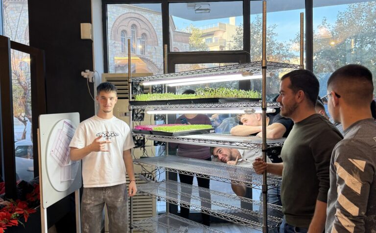 Participants stand beside a microgreens growing rack with lit trays during an EVEC training course in Athens