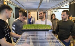 Participants gather around a microgreens growing rack as a tray of microgreens is placed on the shelf, during an EVEC training course in Athens